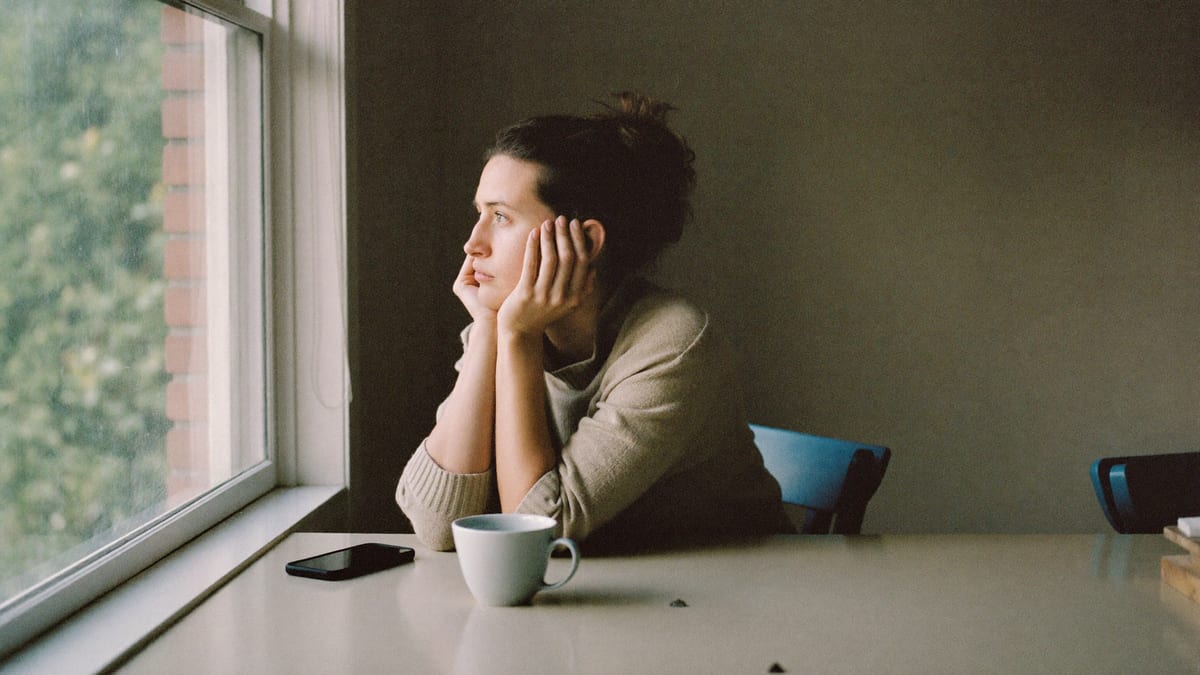A woman sitting at a kitchen table in morning light, looking out of the window, her phone and a mug on the table beside her.